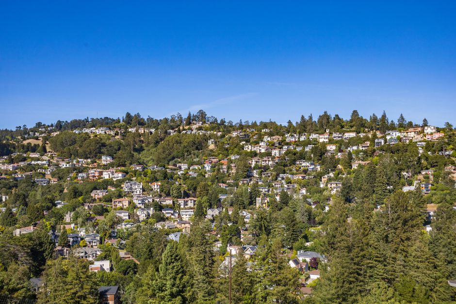 A panoramic view of a densely populated residential area on a hillside, featuring numerous modern detached houses surrounded by lush green trees and vegetation under a clear blue sky. The houses are o