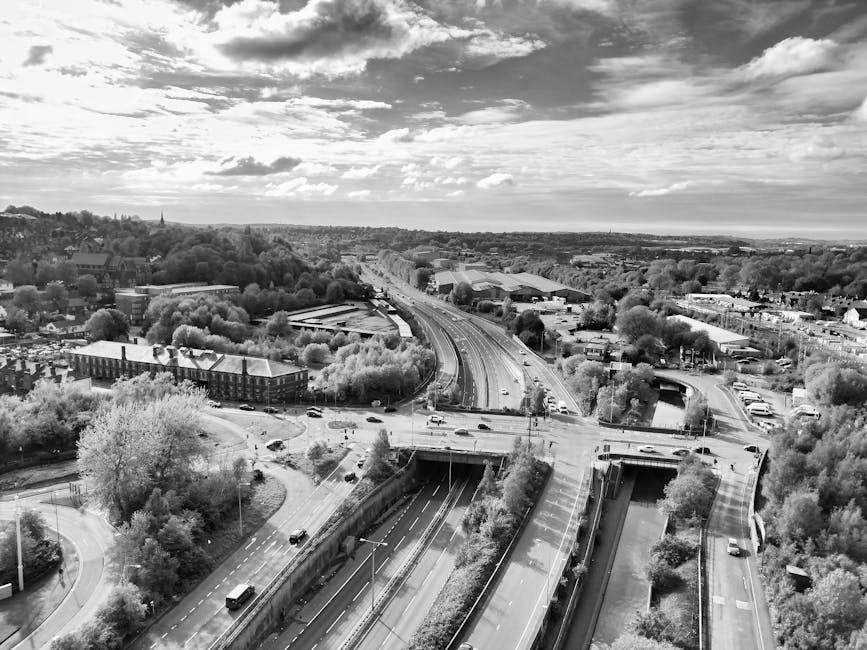 A black and white aerial photograph showing a broad view of Strawberry Hill and Pope's Grove in London, with an extensive road network including a multi-lane motorway running through the scene. The motorway features several vehicles, with some lanes appearing empty, while road bridges and underpasses are visible beneath the elevated sections. Surrounding the roads are patches of greenery, trees, and residential or commercial buildings, with a mixture of dense clusters of trees and open spaces. The sky is partly cloudy with varying cloud formations casting shadows over the landscape. The image captures the urban environment and transportation infrastructure relevant to house removals, illustrating the logistical context of moving services provided by Man with Van Strawberry Hill, particularly during a home relocation process involving furniture transport and clearance across this area.