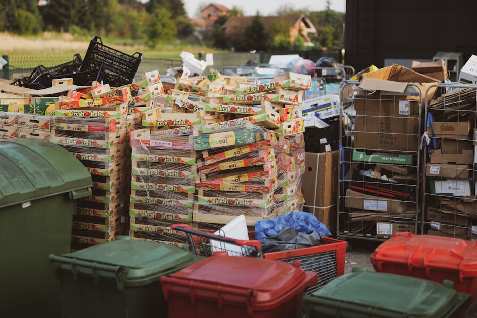 An outdoor scene showing a large stack of flattened cardboard fruit boxes labeled 'fresh fruits' arranged on pallets, ready for disposal or recycling during a house relocation process. Surrounding the boxes are several plastic waste bins in green and red colors, with some open and partially visible. To the right, there are metal trolleys filled with various packing supplies, including cardboard boxes, plastic wrapping, and packaging materials. The background features a paved area near a building doorway or loading zone, with a partially open door and other moving-related items visible, indicating an organized effort to clear bulky waste from a property as part of a home relocation or furniture transport service. The scene is well-lit with natural daylight, and [COMPANY_NAME], Man with Van Strawberry Hill, is involved in the packing and loading process, supporting efficient moving logistics and waste disposal.