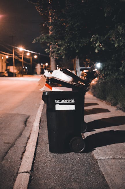 A black wheelie bin positioned on the pavement outside a residential property during nighttime, partially filled with bulky waste items such as cardboard boxes, plastic packaging, and paper waste, some of which are spilling over the top. The bin is marked with the label 'ST. JOHN'S' and is situated on a street illuminated by streetlights, with trees and bushes lining the sidewalk. In the background, a parked vehicle and houses are visible under the evening sky. This scene suggests a waste disposal process associated with house removals or relocating services, as provided by Man with Van Strawberry Hill, supporting home relocation and furniture transport activities.