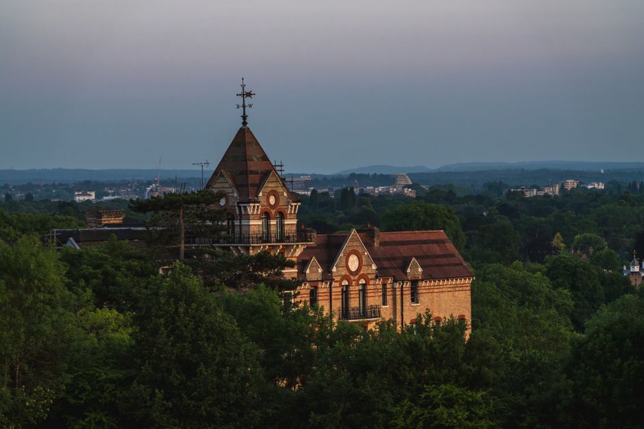 A historic brick church with a pointed steeple topped by a weather vane is visible in the image, surrounded by lush green trees and foliage in Strawberry Hill, Richmond upon Thames. The church has arched windows, decorative brickwork, and a tiled roof, set against a background of distant rolling hills and a partly cloudy sky during daytime. The scene captures the building's prominent position within a residential area, with the church’s structure rising above the treeline. This setting, characteristic of local architecture and landscape, is relevant to home relocation and furniture transport processes as it exemplifies the typical environment in which house removals and logistics are conducted, especially for residents in the area exploring bulk-item or special item rules for moves as per Richmond upon Thames regulations. The natural lighting highlights the details of the brickwork and the surrounding greenery, conveying an accurate visual impression suitable for accessibility and SEO purposes in moving service content for [COMPANY_NAME].