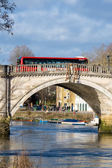 A red double-decker bus is visible crossing a historic stone bridge over a river in Strawberry Hill, with the bridge featuring ornate balustrades and a curved archway. Below, the river reflects the blue sky and nearby trees, some of which are leafless indicating a colder season. In the background, a row of residential buildings and trees line the riverbank. The scene is well-lit with natural daylight, highlighting the architectural details of the bridge and the surrounding environment. This image illustrates a typical urban transport scene relevant for home relocation or furniture transport services by Man with Van Strawberry Hill, set against the backdrop of an outdoor moving logistics context.