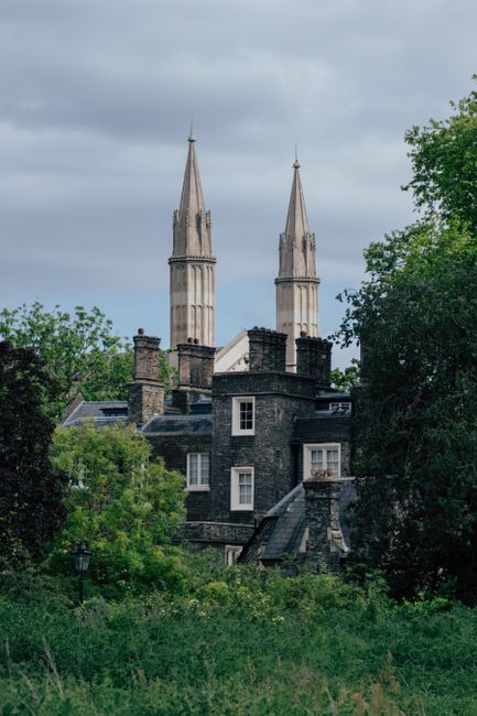 A historic brick church with a pointed steeple topped by a weather vane is visible in the image, surrounded by lush green trees and foliage in Strawberry Hill, Richmond upon Thames. The church has arched windows, decorative brickwork, and a tiled roof, set against a background of distant rolling hills and a partly cloudy sky during daytime. The scene captures the building's prominent position within a residential area, with the church’s structure rising above the treeline. This setting, characteristic of local architecture and landscape, is relevant to home relocation and furniture transport processes as it exemplifies the typical environment in which house removals and logistics are conducted, especially for residents in the area exploring bulk-item or special item rules for moves as per Richmond upon Thames regulations. The natural lighting highlights the details of the brickwork and the surrounding greenery, conveying an accurate visual impression suitable for accessibility and SEO purposes in moving service content for [COMPANY_NAME].
