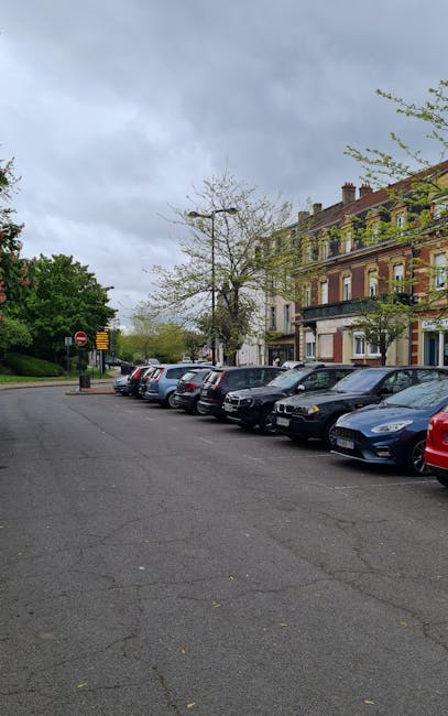 A row of parked cars lining a street in Strawberry Hill, with a sidewalk and residential buildings in the background. The street appears to be relatively narrow, with some trees and street signs visible. The sky is overcast, suggesting cloudy weather. This setting relates to house removals and relocation services, as parking limitations and street access can present challenges for professional movers such as Man with Van Strawberry Hill during home relocation or furniture transport in residential areas.