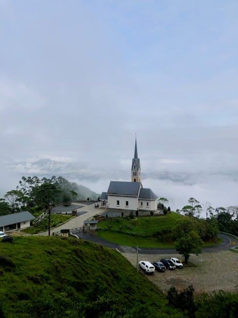 A scenic view of a white church with a tall, pointed steeple situated on a grassy hilltop surrounded by trees and a winding driveway. Several parked cars are visible near the church, with a few people standing around, possibly involved in packing or preparing for home relocation. The background shows misty clouds and fog partially covering the landscape, creating a serene and atmospheric setting. The scene is outdoors on a cloudy day, and the overall environment suggests a rural or semi-rural area, which could be relevant in the context of house removals or moving services like those offered by Man with Van Strawberry Hill.