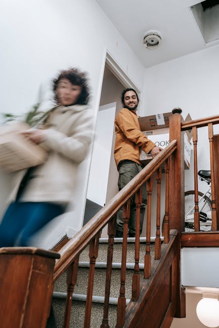 Inside a home during a house removal, a man from Man with Van Strawberry Hill is seen on a staircase holding a cardboard box filled with smaller packages, preparing for furniture transport as part of a home relocation. The staircase features wooden banisters and carpeted steps, with the man smiling and wearing casual work clothing. A second individual, partially visible and slightly blurred, is carrying another box towards the doorway, indicating active packing and moving efforts. The interior lighting is natural, and other boxes are visible on the landing, some wrapped in plastic or packing materials. A bicycle is partially visible in the background, suggesting the belongings being moved include various household items. This scene depicts a typical loading process during a professional removals service, with furniture and boxes positioned for efficient transport to a moving vehicle, illustrating the packing and logistical stages involved in house removals carried out by Man with Van Strawberry Hill.