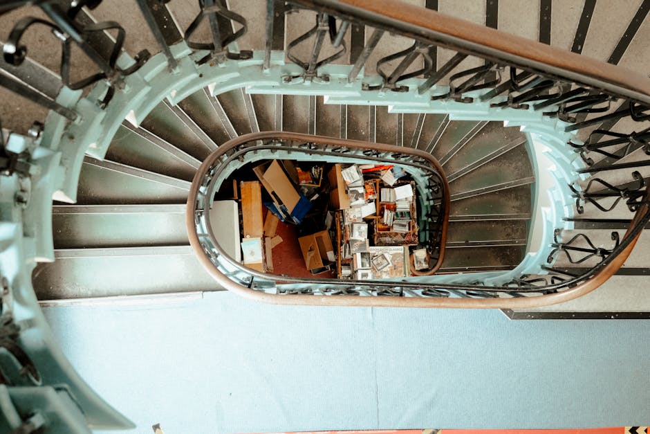 Two movers from Man with Van Strawberry Hill are carefully carrying a large green upholstered armchair down a narrow indoor staircase during a home relocation. The staircase is carpeted with dark brown runners and has black metal handrails on both sides. The scene is well-lit with natural light coming from a window at the bottom of the stairs, revealing exposed brickwork around the window frame. One mover, wearing a black jacket and red shoes, is positioned at the front, gripping the armchair securely, while the second mover, wearing a dark jacket and a headband, supports the opposite side. Cardboard padding and plastic wraps are partially visible, indicating packing materials used for furniture protection during transport. The setting suggests a professional moving process involving loading and furniture transport within a residential property, typical of services offered by Man with Van Strawberry Hill.