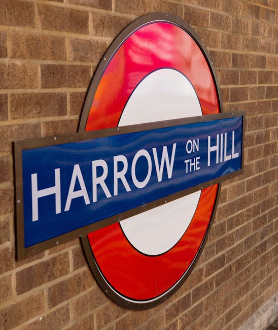 A close-up image of the Harrow on the Hill London Underground station sign mounted on a brick wall. The sign features a circular red and white tube emblem with a rectangular blue plaque across the middle displaying the station name in white capital letters. The plaque partially covers the circular emblem and is positioned at an angle, suggesting the sign has been temporarily removed or is being relocated. The background consists of brown brickwork, and the lighting appears natural, highlighting the textures of both the sign and the brick wall. This setting relates to house removals and moving logistics, especially when considering transportation and station access during home relocation projects, consistent with services offered by Man with Van Strawberry Hill.