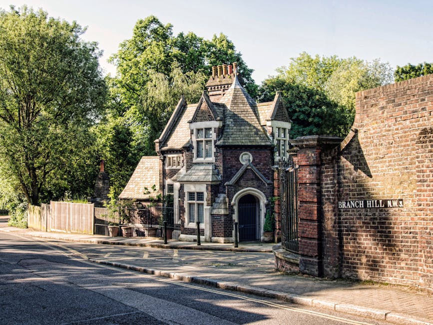 A narrow residential street with a paved road and tiled sidewalk, featuring a historic, Victorian-style house with a steeply pitched slate roof, ornate chimney stacks, and bay windows enframed by decorative stonework. The house is located behind a black wrought iron gate and fence, with a small garden and trees providing greenery around the property. To the right, a red brick wall with a street sign reading 'Branch Hill N.W.3' marks the boundary of the property. A tall, leafy tree towers behind the house, casting dappled shadows on the street and building. The scene is illuminated by bright daylight, clearly showing the textures of brick, slate, and foliage. Occasionally, [COMPANY_NAME] may assist in home relocation or furniture transport, involving careful packing and loading processes, as seen in the environment's organized and secure setting.