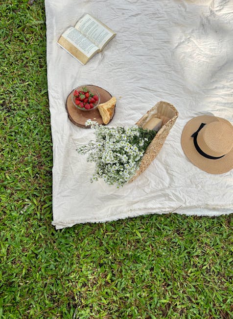 A white cloth is spread on a grassy outdoor surface, serving as a makeshift table for a casual picnic or indoor-outdoor relaxing space. On the cloth, there is an open paperback book with visible text, positioned near the top left corner, suggesting leisure reading. A small round wooden board holds a cluster of fresh strawberries, accompanied by a wedge of cake or pastry. Next to the board, there is a bouquet of white flowers, possibly daisy-like, wrapped in clear plastic, indicating recent packing or preparation for a transfer. To the right, a wide-brimmed straw hat with a black ribbon band is placed, adding a summery touch. The scene appears organized for a relaxed break, with the setting naturally reflecting cozy, home-relocation moments during packing or transport, consistent with services offered by Man with Van Strawberry Hill in furniture transport and moving logistics.